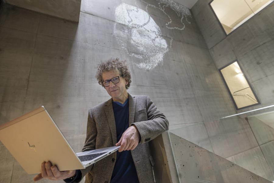 Mies seisoo tietokone kädessään, hänen takana on korkea seinä missä digitaalinen taideteos näkyy
